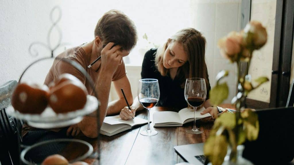 A man and a woman sit at a table, writing in notebooks and reading books.
