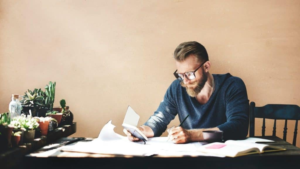A man with a beard and glasses sits at a desk, writing and holding a calculator.