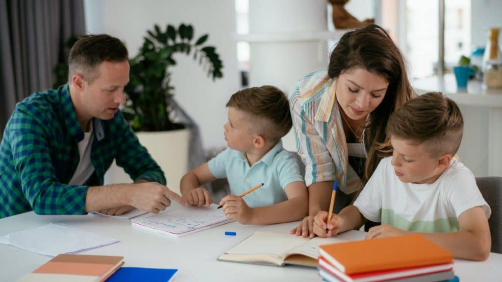 A couple helps two young boys with their homework at a table.