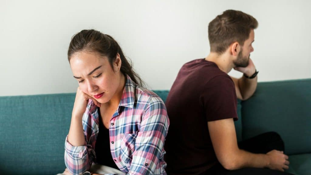 A sad-looking woman sits on a couch while a man behind her faces away.