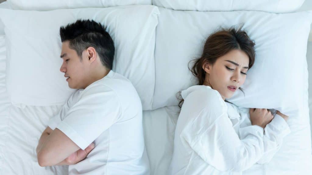 A man and a woman lie in bed facing away from each other, looking unhappy.