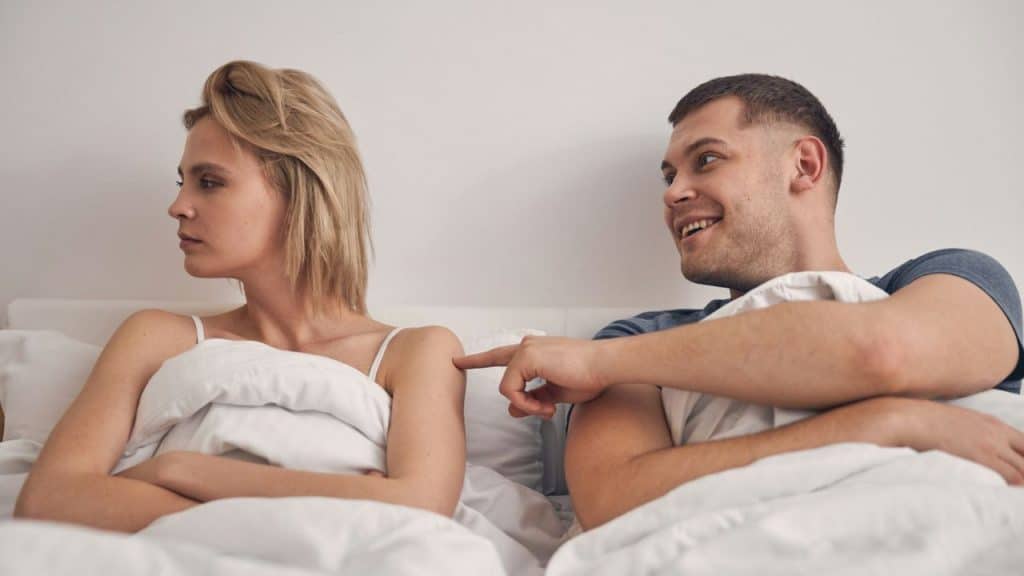 A man in bed smiles and points at a woman in bed who looks away from him.