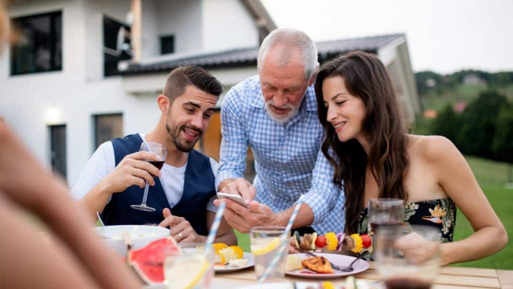 A group of three adults at an outdoor table look at a phone together.