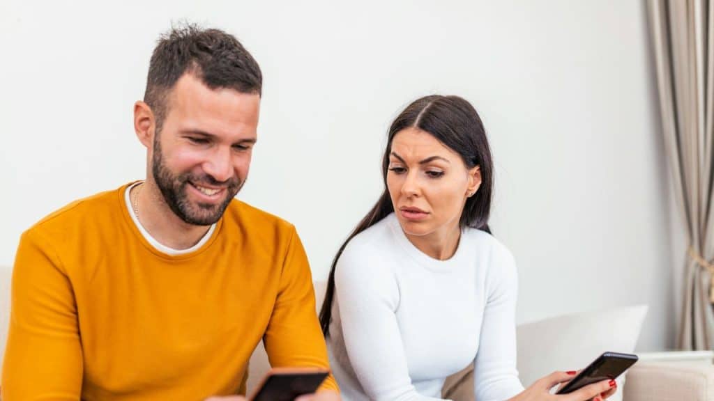 A man smiles at his phone while a woman beside him looks at her phone with concern.