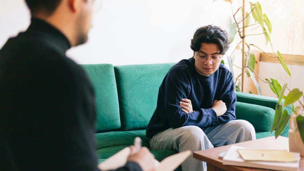 A person with glasses sits on a green couch, looking down with arms crossed.