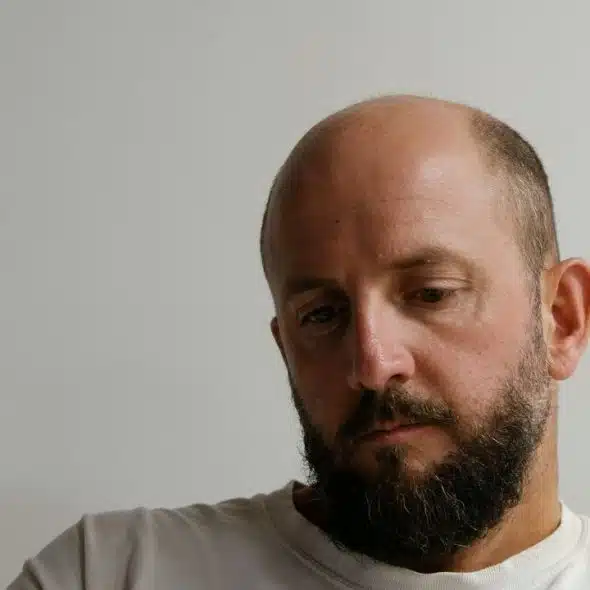 A close-up of a bald man with a beard, looking down thoughtfully.