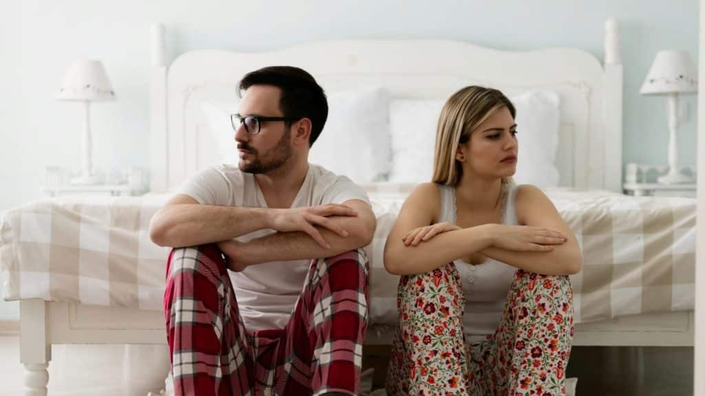 A man and a woman sit on the floor, backs to each other, looking upset.