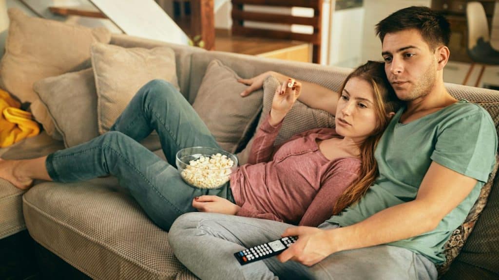 A couple sits on a couch, with the man holding a remote and the woman eating popcorn.