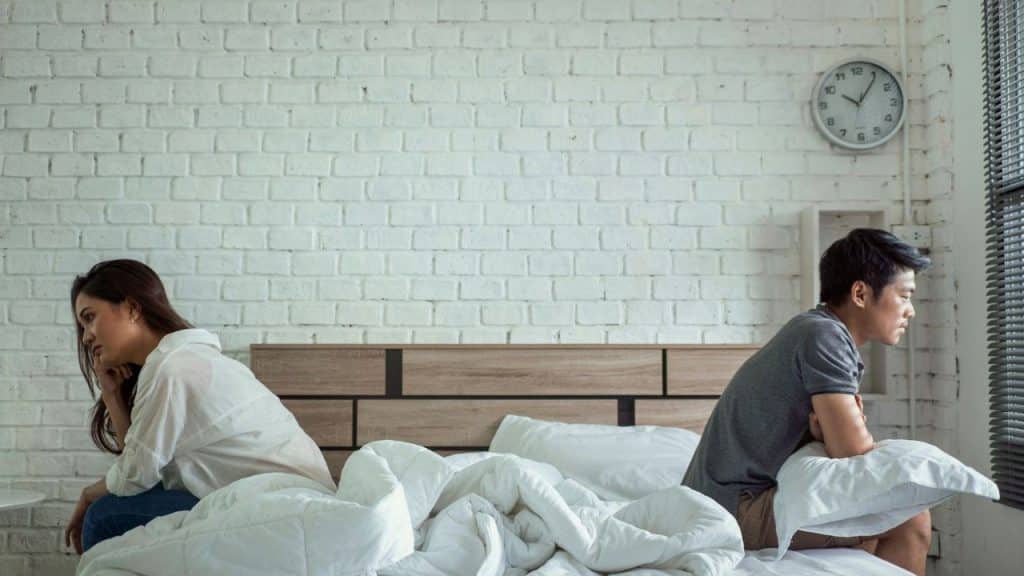 A man and a woman sit on a bed with their backs to each other, looking upset.