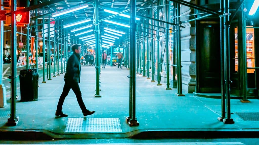 A man walks under a scaffolding tunnel lit with blue fluorescent lights at night.