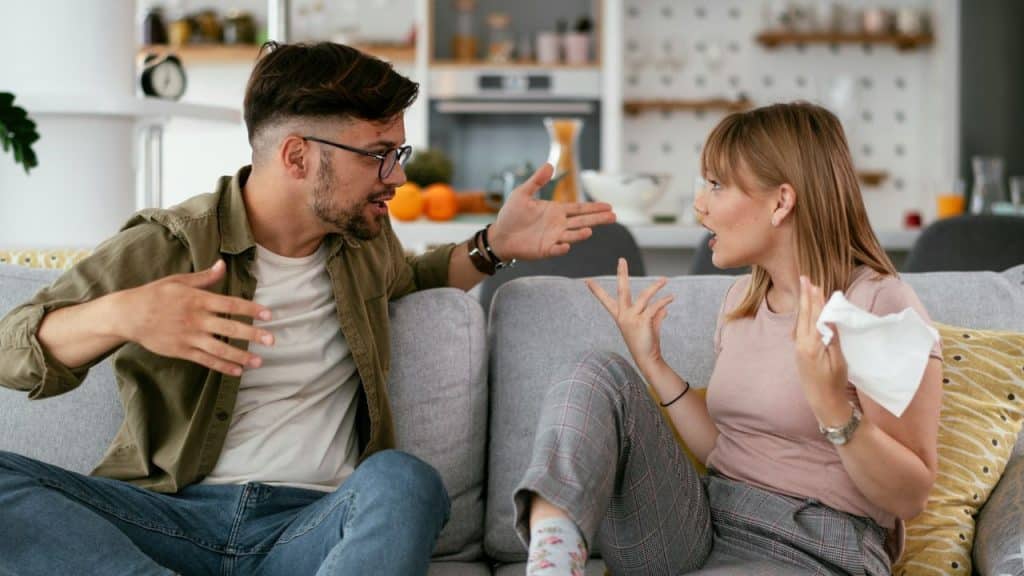 A man and woman sit on a couch, gesturing with their hands as they argue.