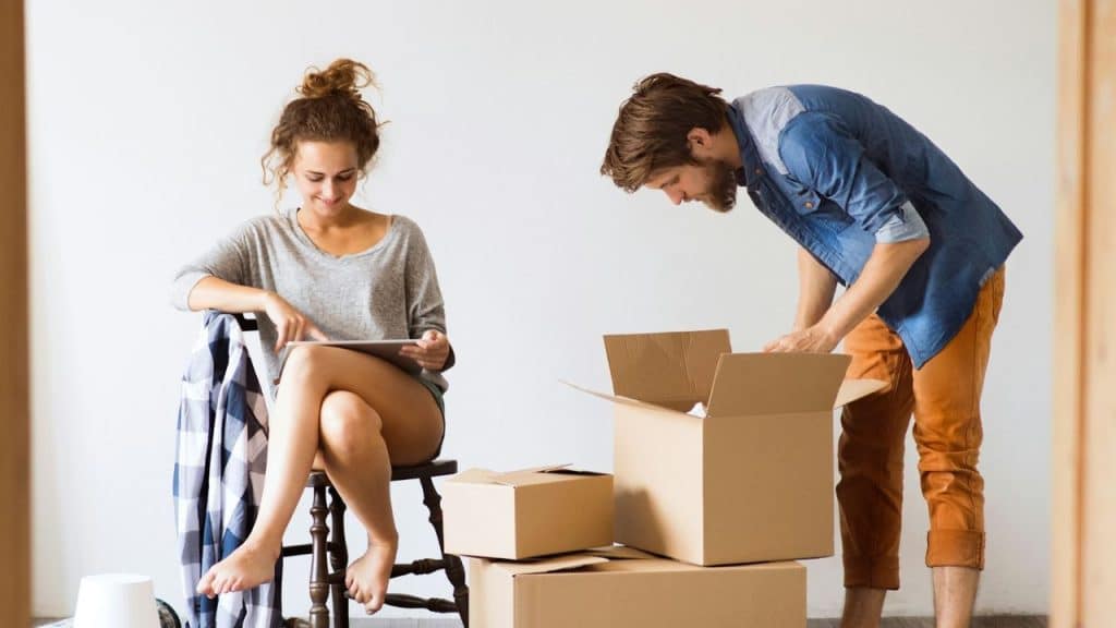 A woman sits and uses a tablet while a man unpacks a box nearby.