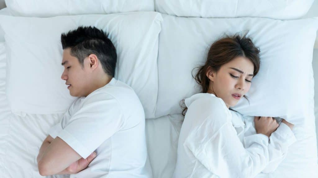 A man and a woman lie in bed facing away from each other, looking unhappy.