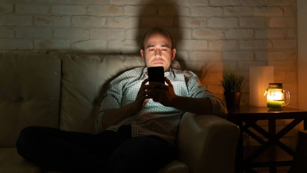 A man sits on a couch in a dimly lit room, looking at his phone.
