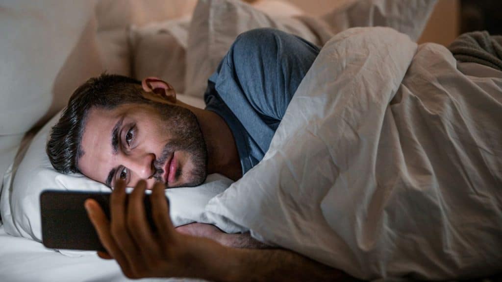 A serious man is lying in bed, looking at his smartphone at night.