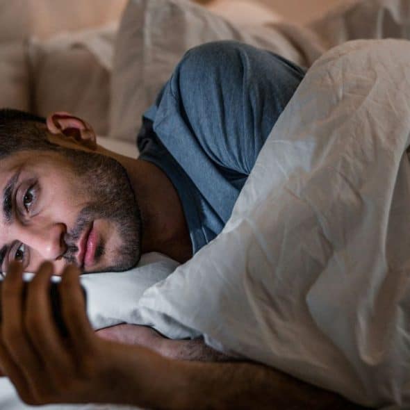 A serious man is lying in bed, looking at his smartphone at night.