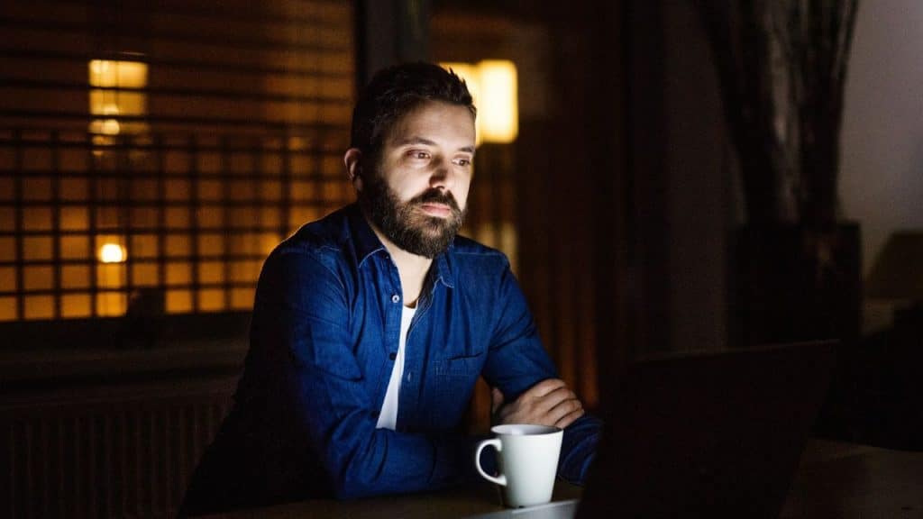A tired man with a beard sits at a table, looking at a laptop.