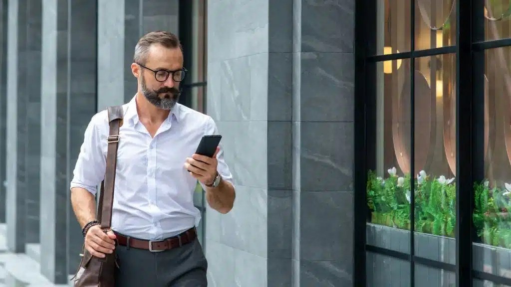A well-dressed, bearded man with glasses is walking while looking at his phone.
