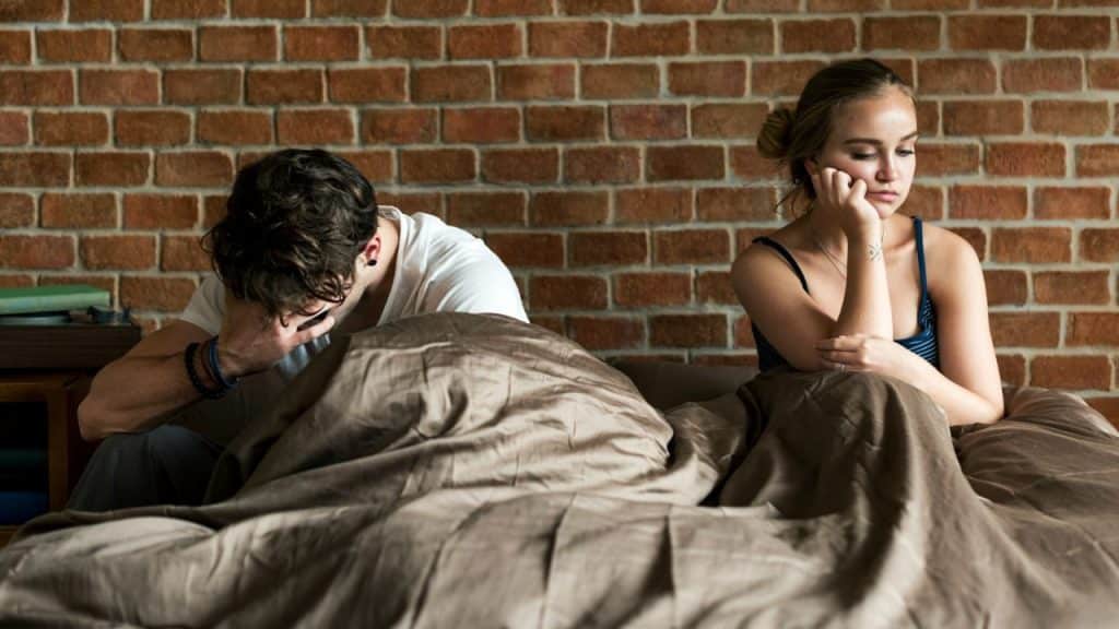 A man and woman sit on a bed with their backs to each other, looking upset.