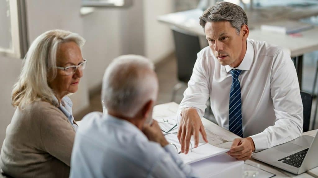 A middle-aged man in a white shirt and tie explains a document to an older couple.