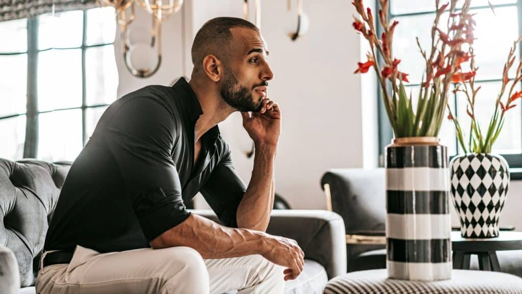A bearded man in a black shirt sits on a couch, looking away thoughtfully.