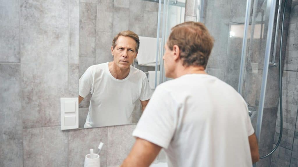 A man in a white t-shirt looks at his reflection in a bathroom mirror.