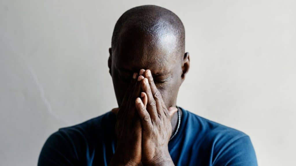 A sad man with his hands over his face looks down.