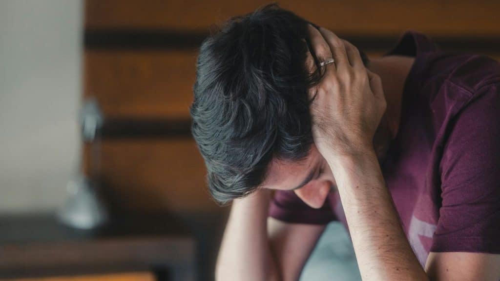 A man with dark, curly hair sits with his hands grasping his head, looking down.