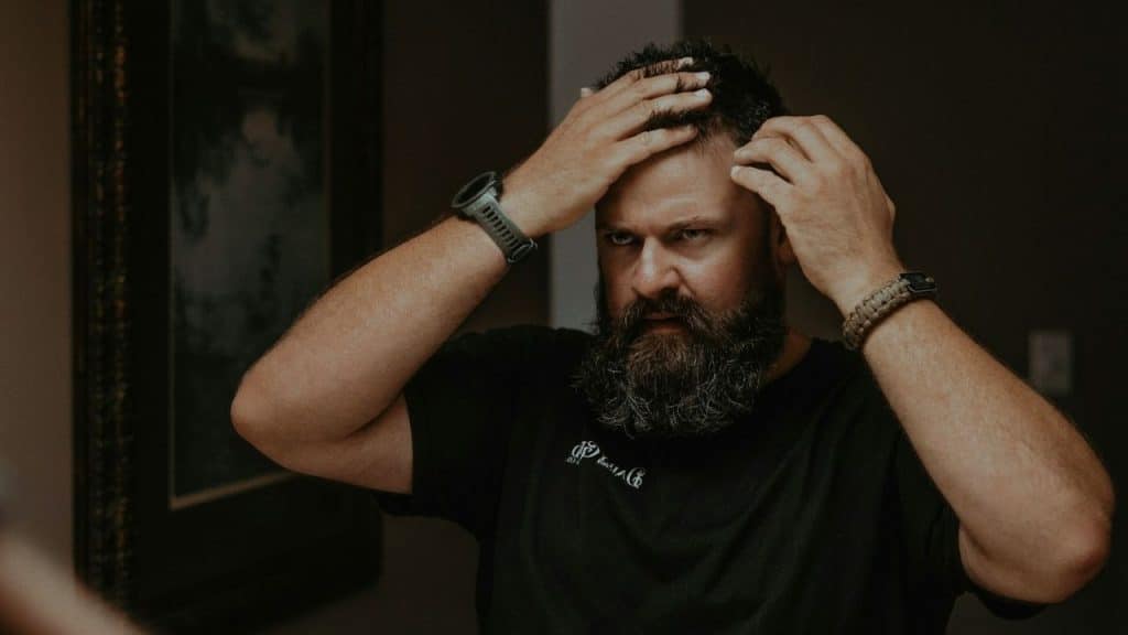 A man with a full beard and dark hair looks in the mirror and checks his hairline.