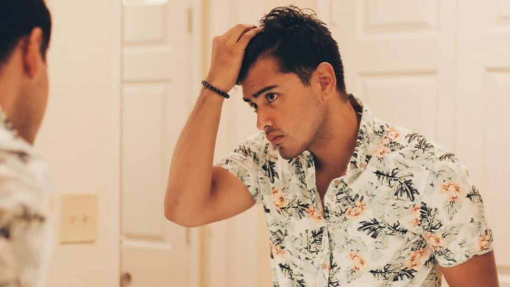A man in a floral shirt looks at his hair in the mirror.