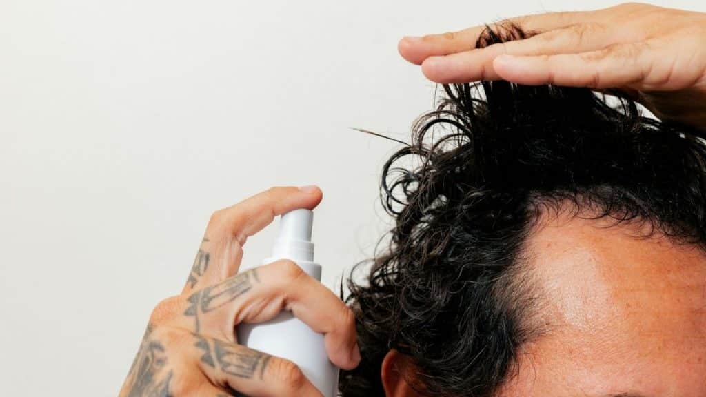 A tattooed hand holds a spray bottle and applies a product to a man's hair.