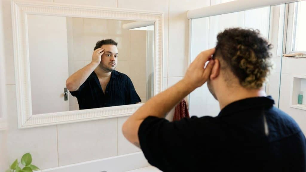 A man with dark curly hair looks at his reflection in a bathroom mirror and checks his hairline.