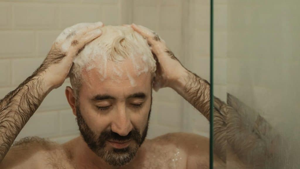 A man with a beard and bleached hair washes his hair with shampoo in the shower.