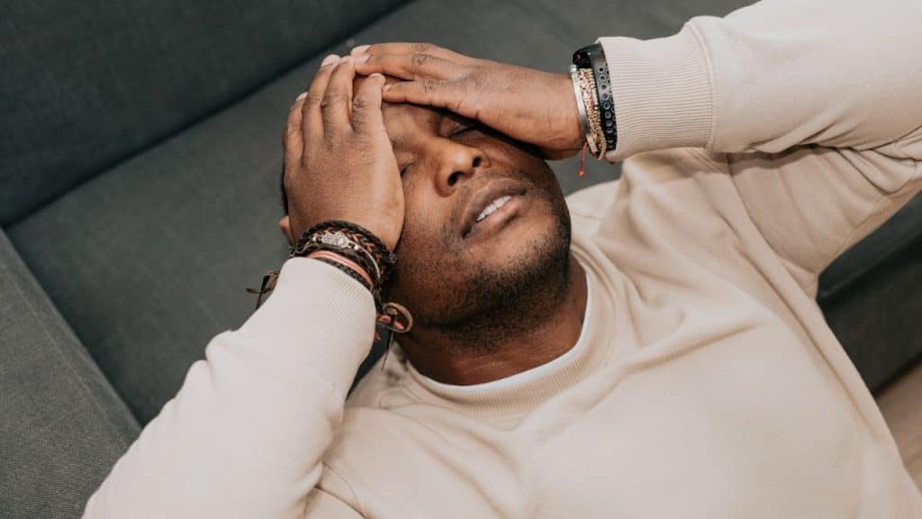 A man lies on a couch with his hands over his face, looking stressed.