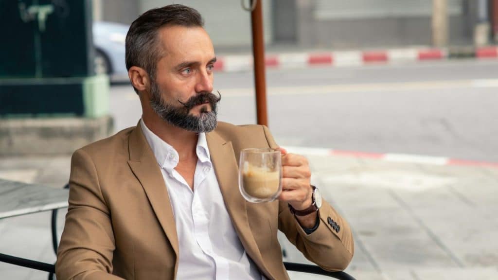 A stylish, handsome, middle-aged man with a full beard and mustache sits at an outdoor cafe.