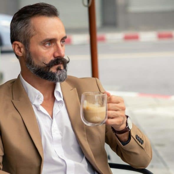 A stylish, handsome, middle-aged man with a full beard and mustache sits at an outdoor cafe.