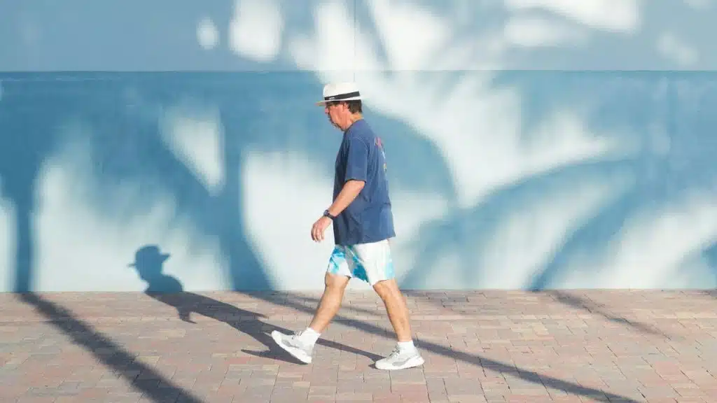 A man in a hat, t-shirt, and shorts walks along a sidewalk with palm frond shadows.