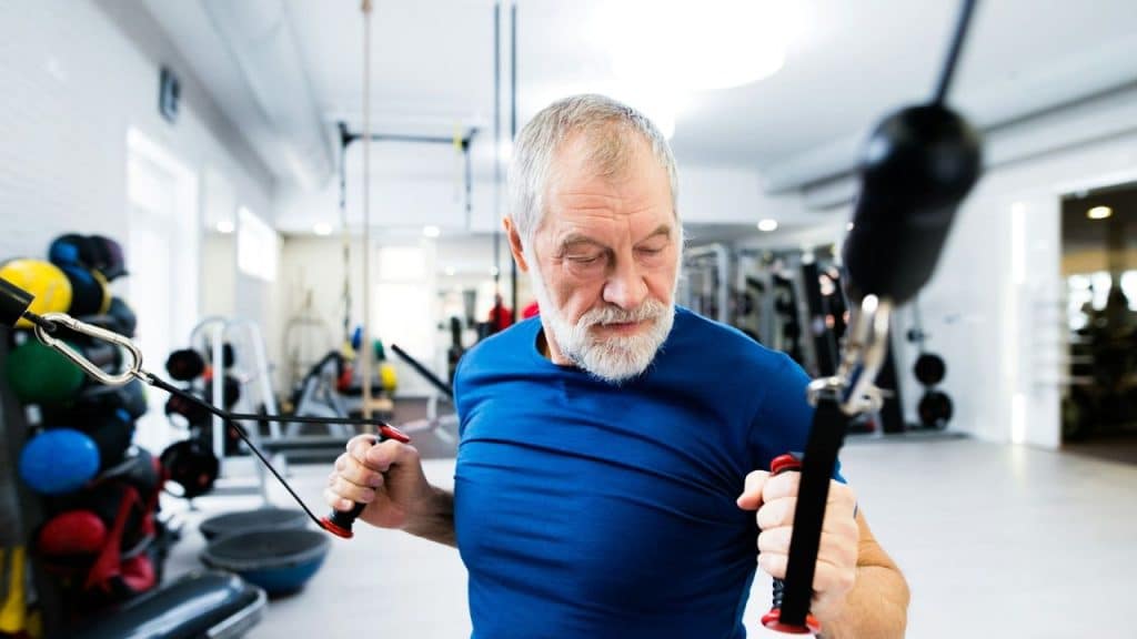 A fit, older man with a beard and gray hair works out with a cable machine in a gym.