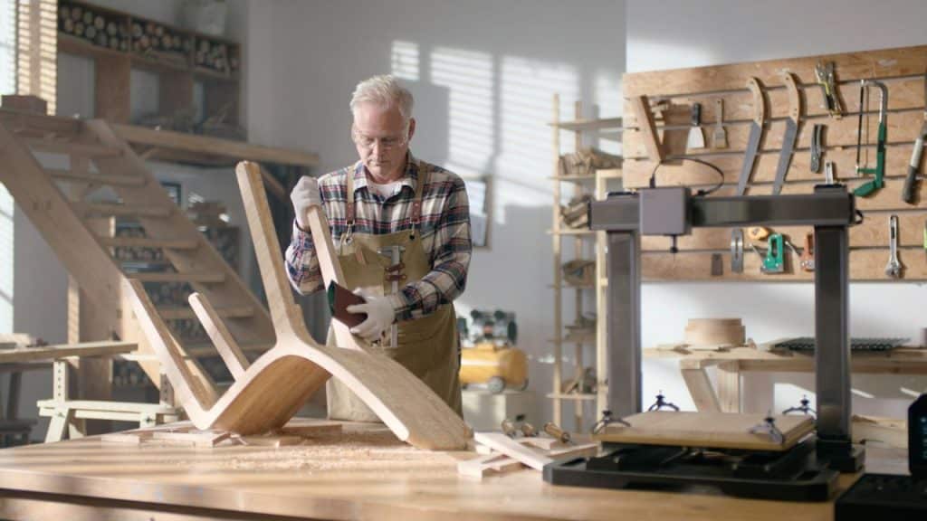 A senior man wearing safety glasses and an apron stands in a workshop and works on a wooden chair.