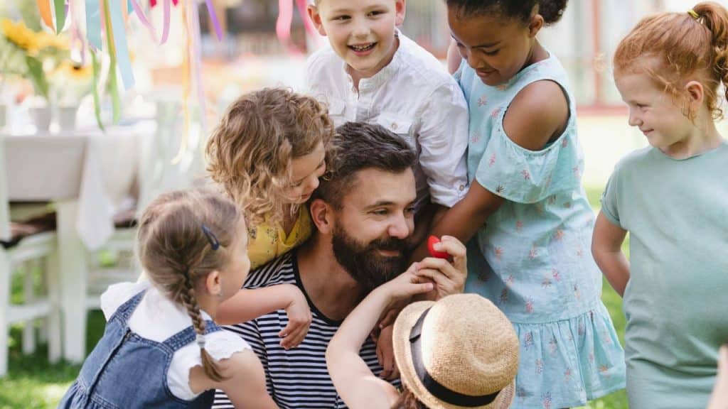 A happy man with a beard sits on the grass and is surrounded by five children.