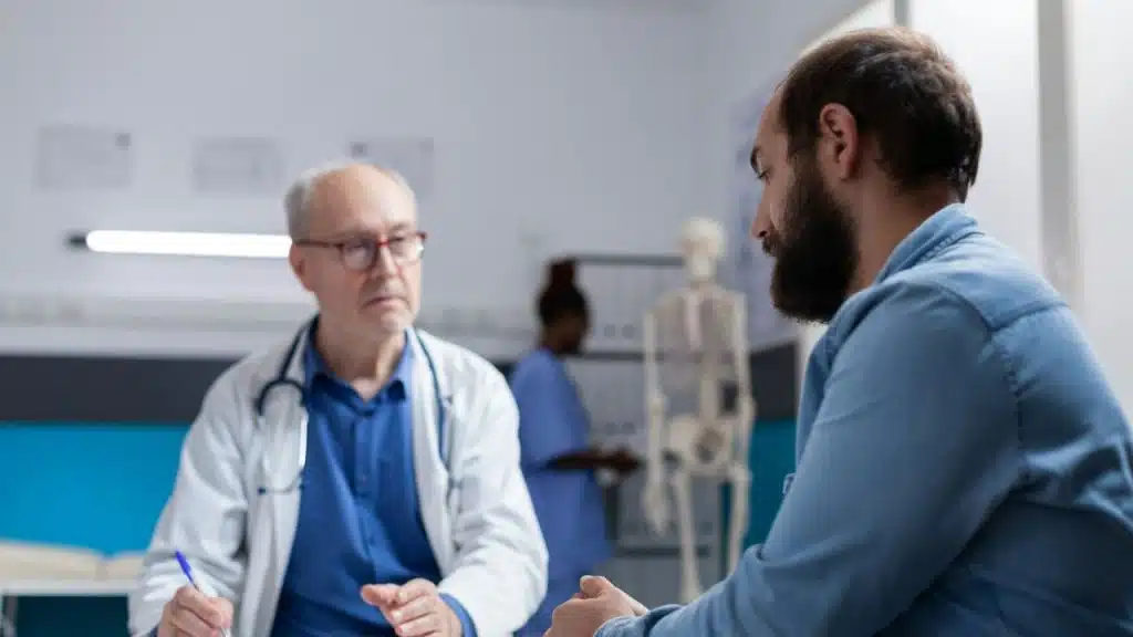 A man with a beard sits opposite a male doctor in a white lab coat.