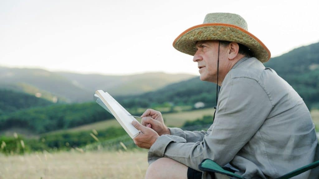 A middle-aged man in a straw hat sits in a field, holding a pencil and notebook.