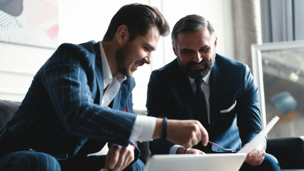 Two handsome men in suits sit together, with one pointing at a laptop screen.