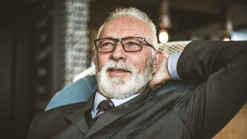 A smiling, gray-haired man in a suit and glasses rests his head in his hands.