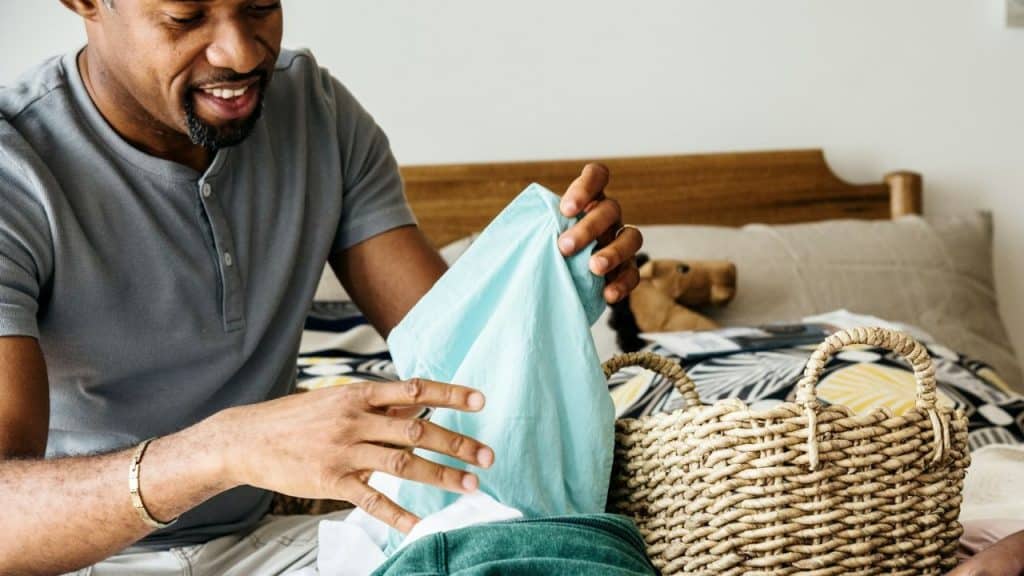 A man is folding a light blue shirt and putting it into a wicker basket.