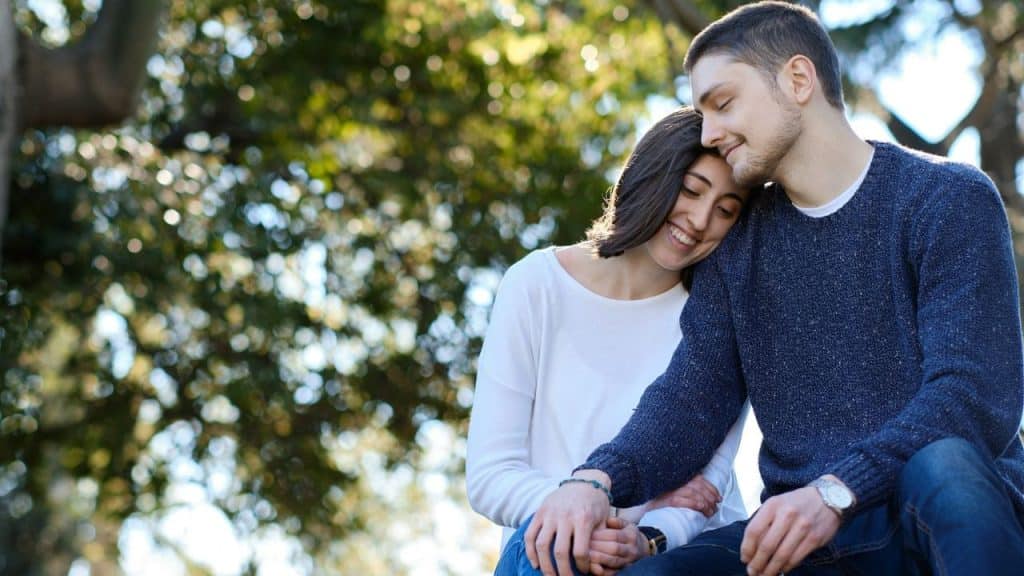 A couple sits close together outdoors, holding hands and smiling.