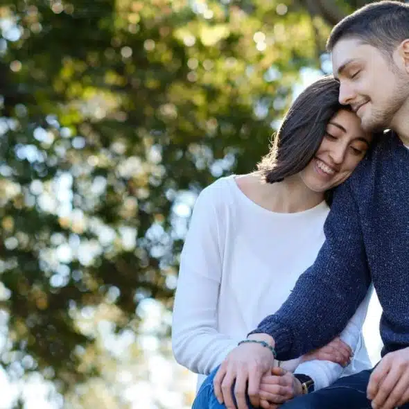 A couple sits close together outdoors, holding hands and smiling.