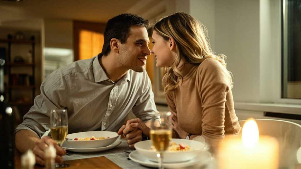 A smiling couple looks at each other at a candlelit table with food and wine.