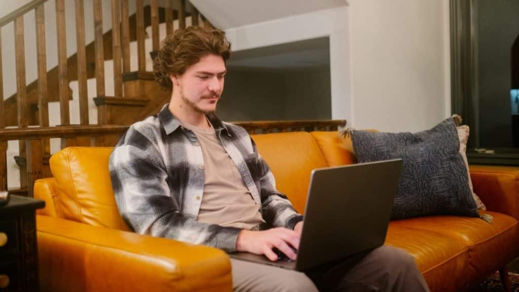 A man with a mustache sits on an orange couch and uses a laptop.