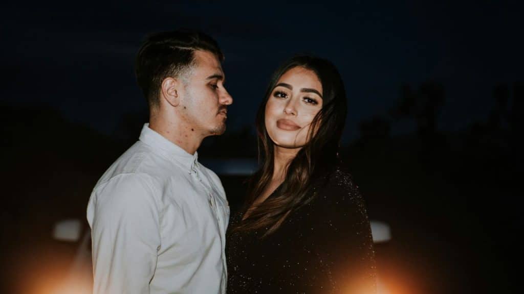 A smiling couple stands close together at night with a bright light in the foreground.